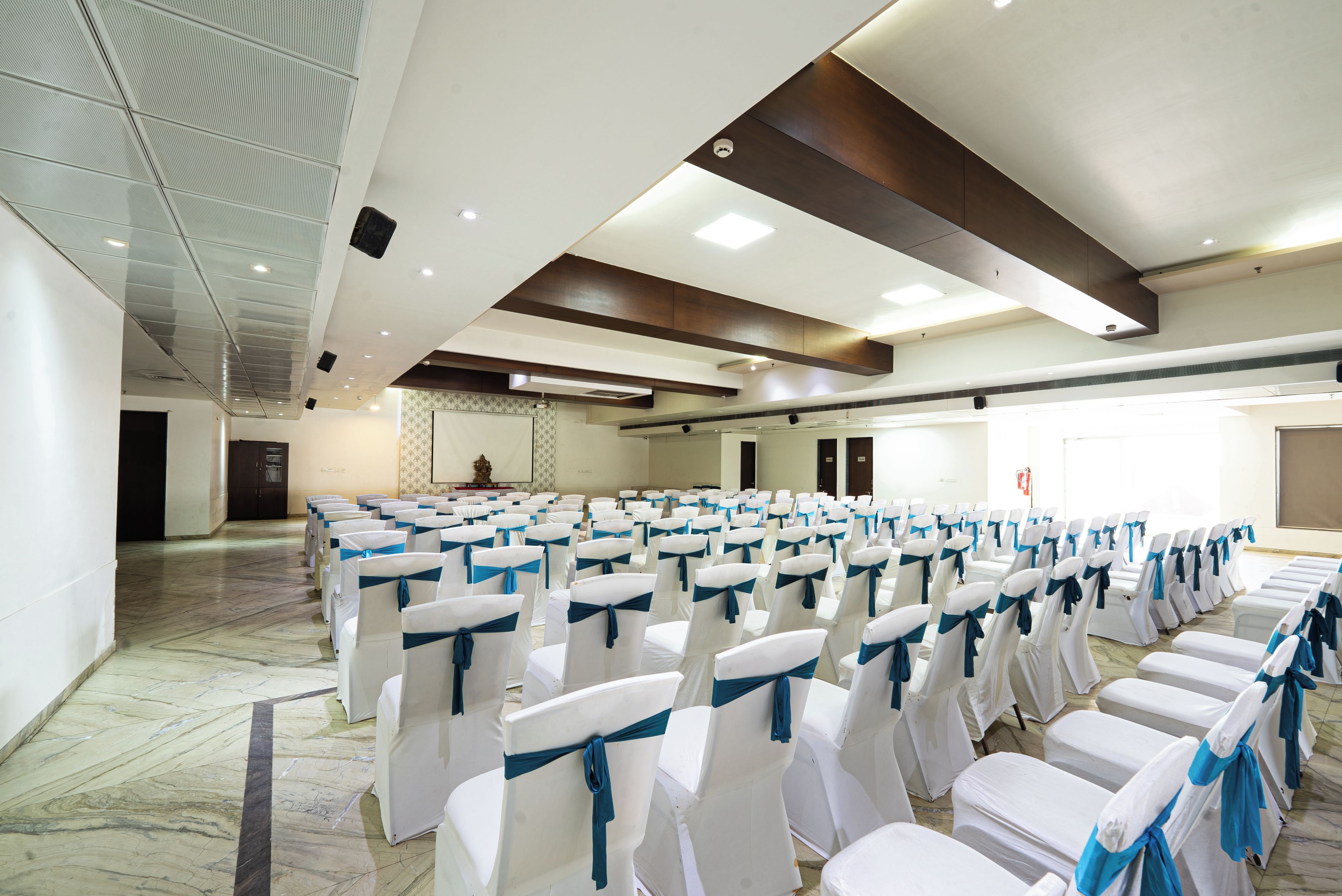 Un salón de conferencias con filas de sillas blancas atadas con cintas azules, piso de mármol y un techo alto con iluminación empotrada.