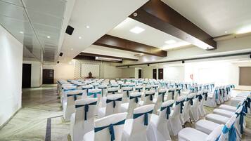 Un salón de conferencias con filas de sillas blancas atadas con cintas azules, piso de mármol y un techo alto con iluminación empotrada.