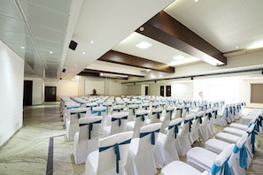 A conference room with rows of white chairs tied with blue sashes, a marble floor, and a high ceiling with recessed lighting. - Manish Aura Grand (Vadodara)