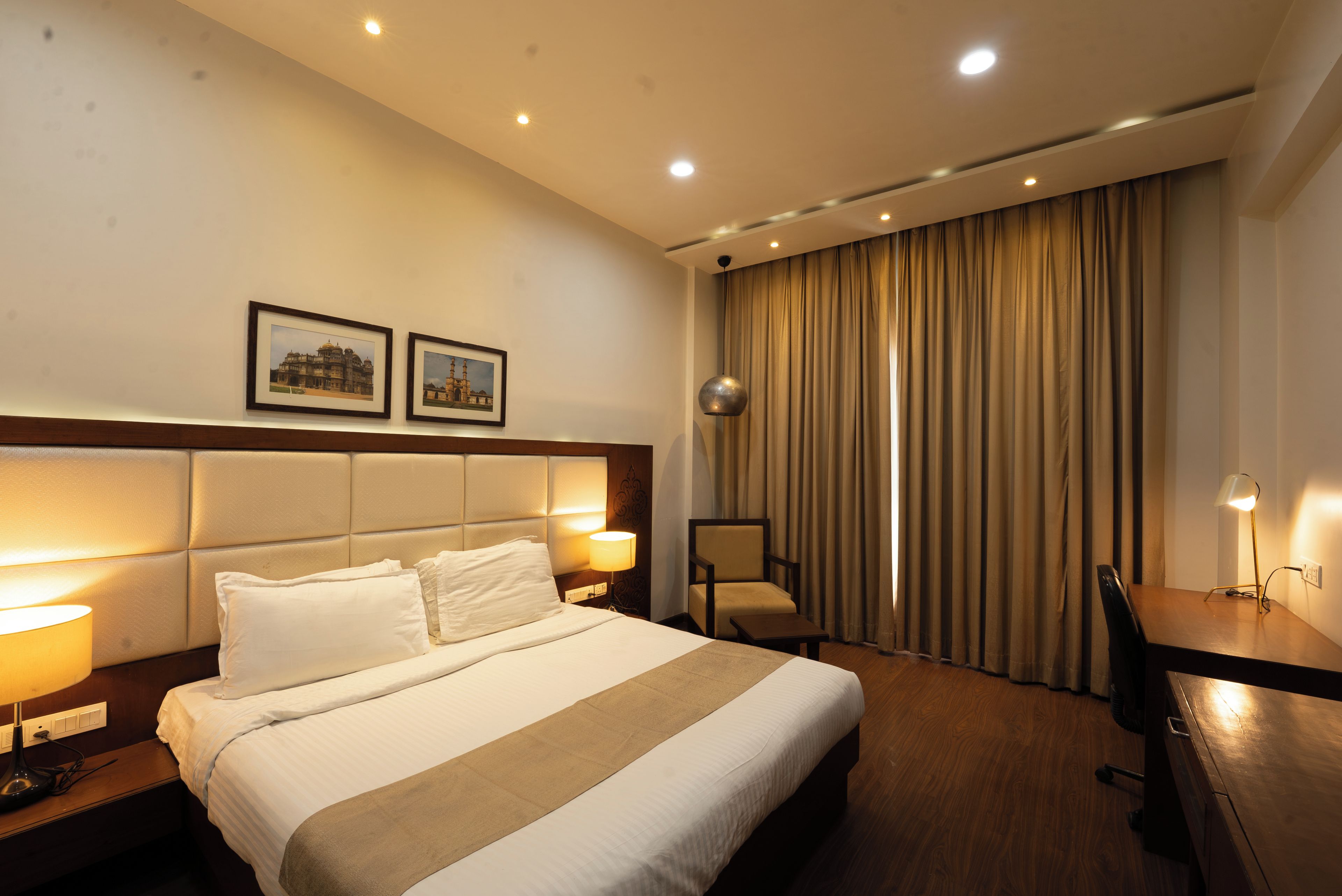 A hotel room with a large bed, a desk, a chair, and two framed pictures on the wall.