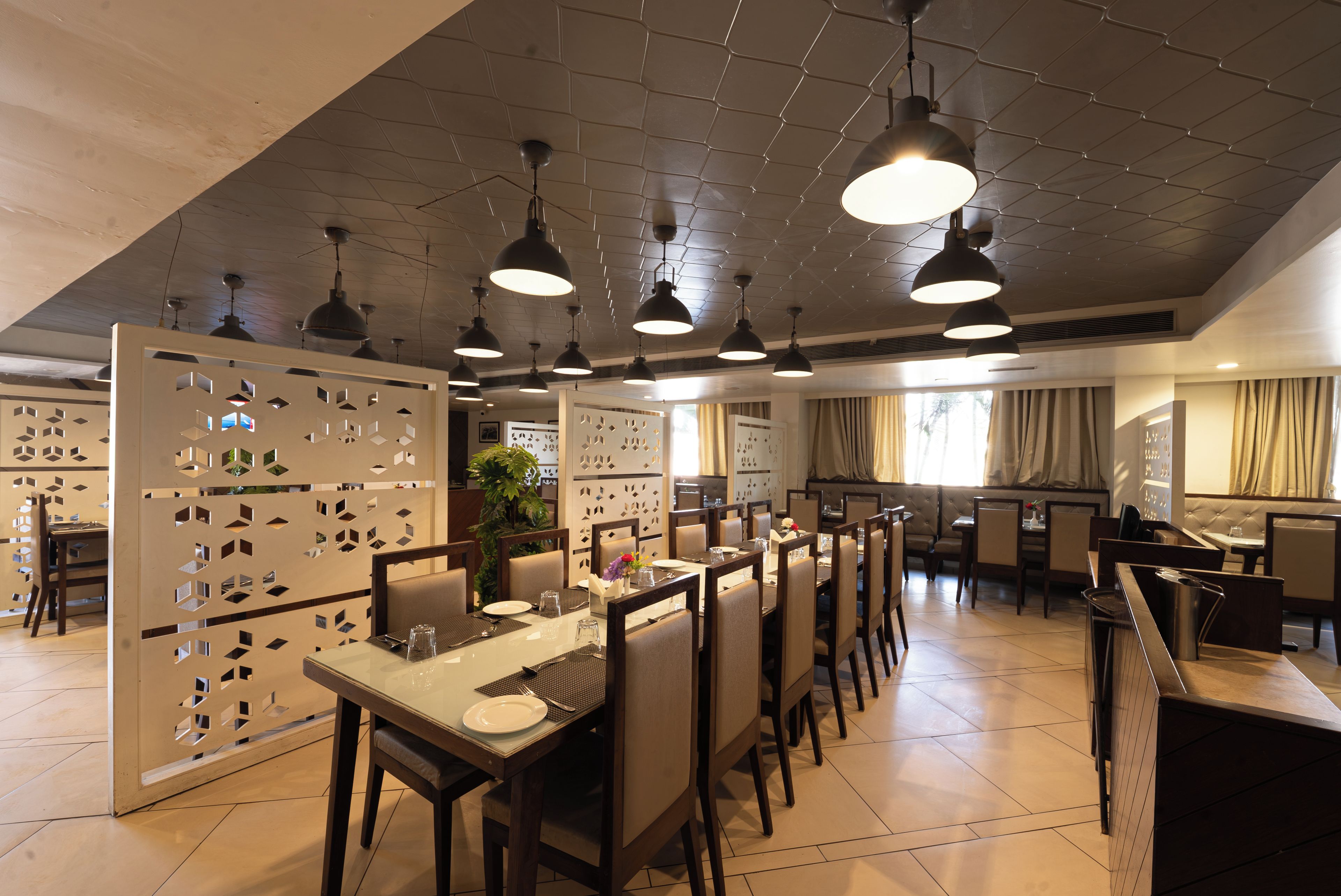 A restaurant with a modern interior, featuring a long dining table with chairs, pendant lighting, and a decorative partition with geometric cutouts.