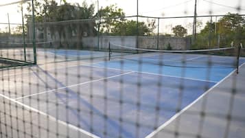 Una cancha de tenis exterior con red, rodeada por una valla y árboles al fondo.