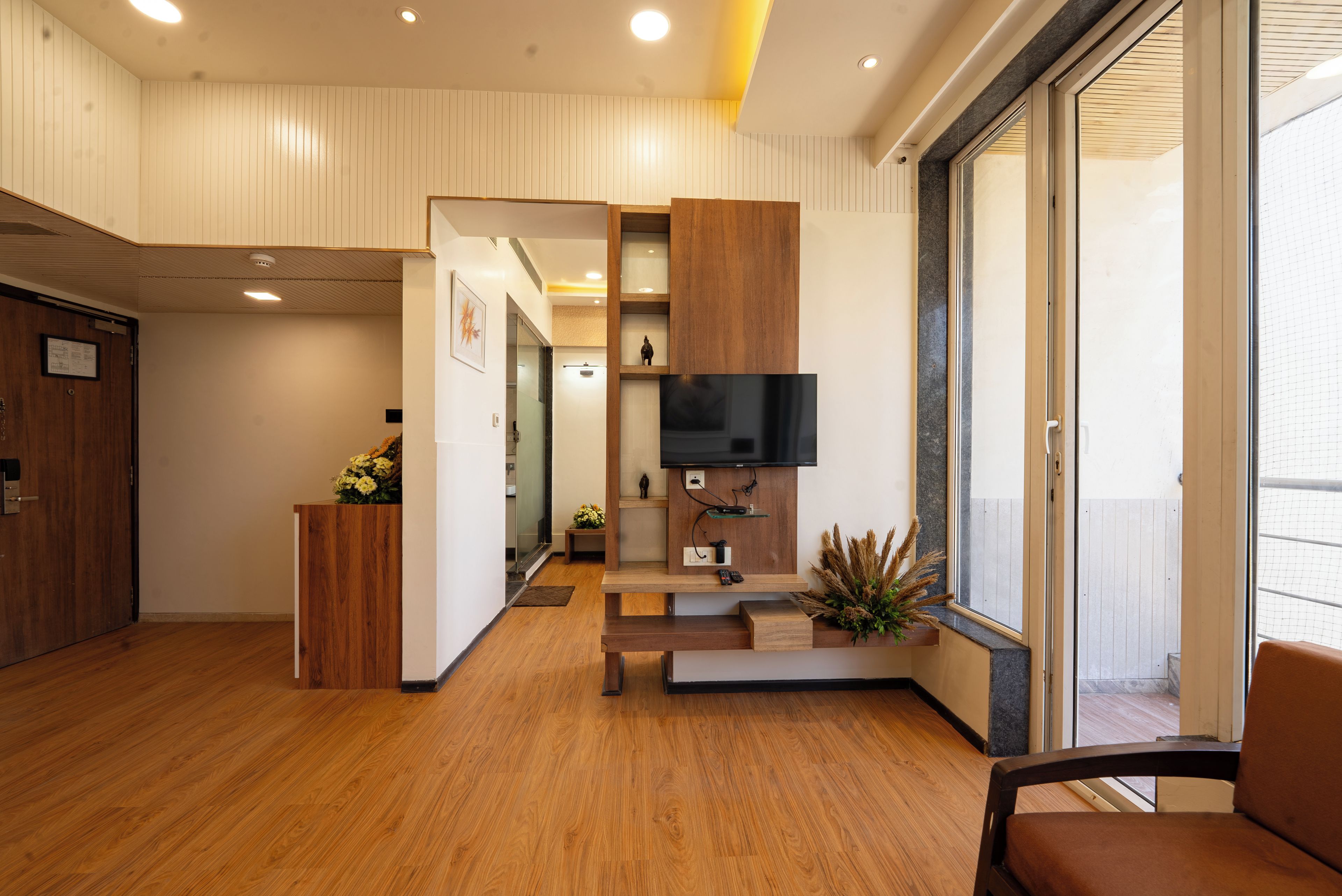 Un interior moderno con suelo de madera, un televisor de pantalla plana montado en un panel de madera, una mesita de madera con un jarrón de flores y un ventanal con vistas.