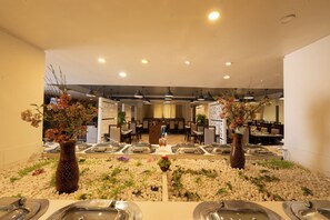 A hotel dining area with buffet service, tables, chairs, and decorative plants. - Manish Aura Grand (Vadodara)