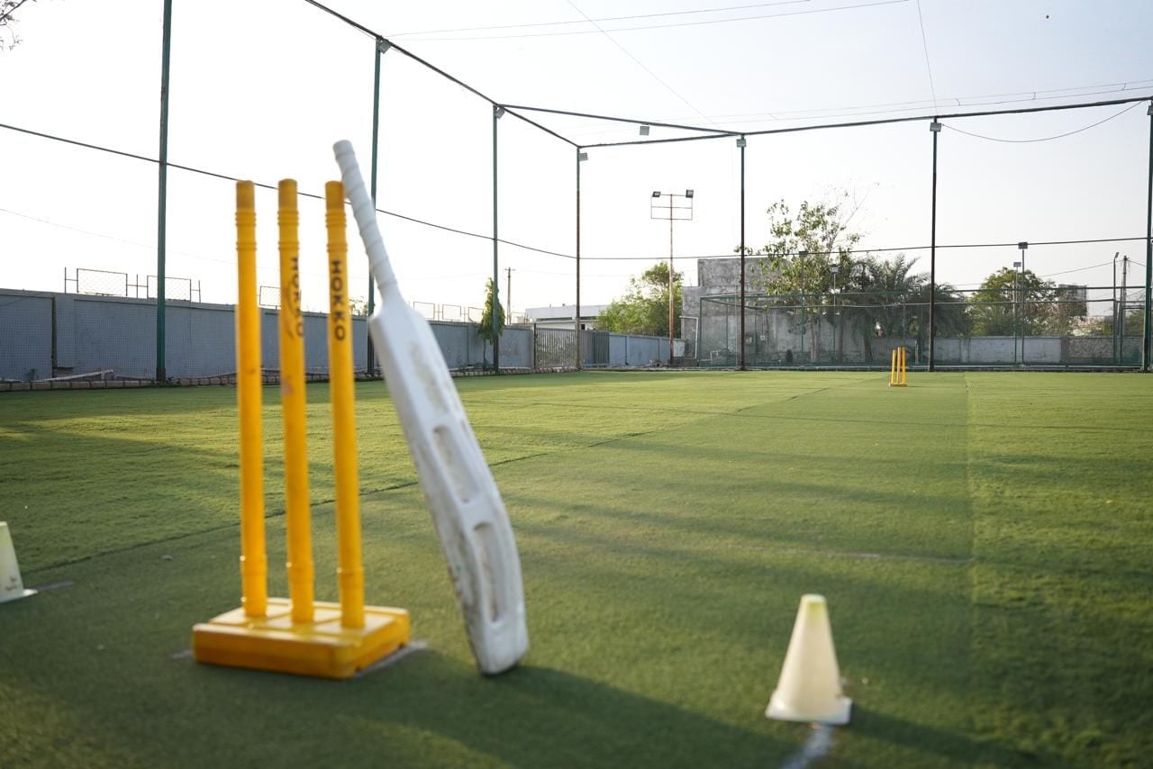 Een cricketveld met wickets, kepers en een omheining, onder een heldere lucht.