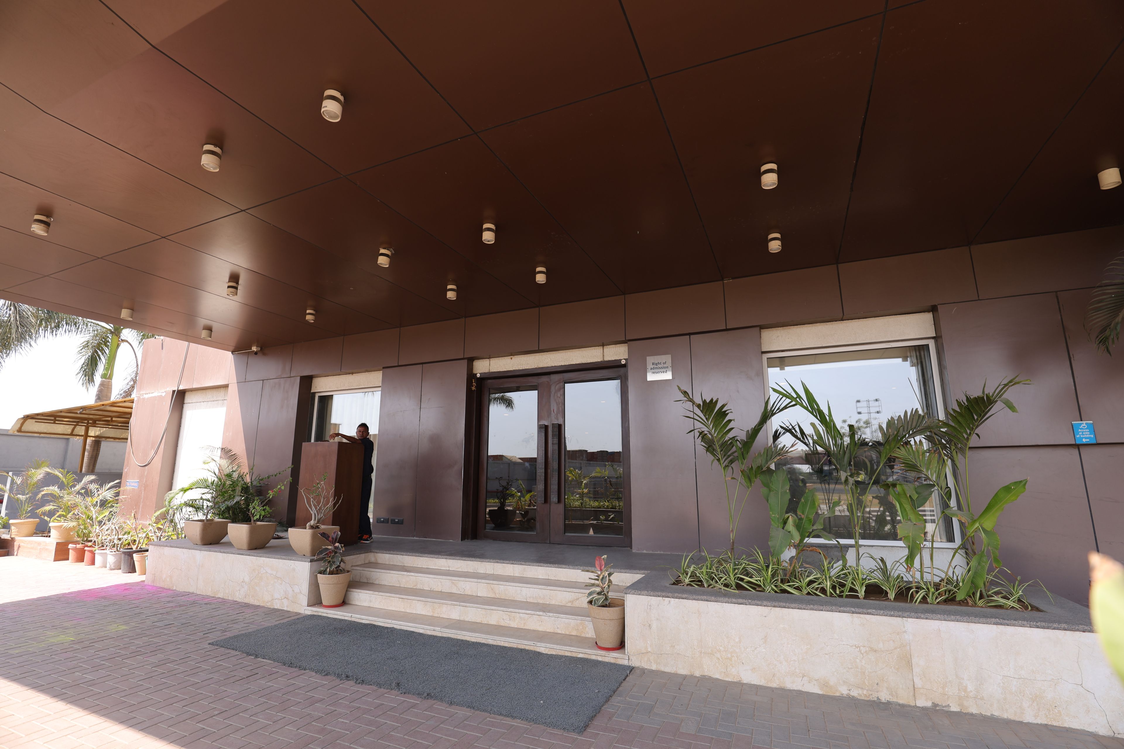 A building entrance with a modern design, featuring a set of steps leading to a glass door, potted plants, and a clear sky visible through the windows.