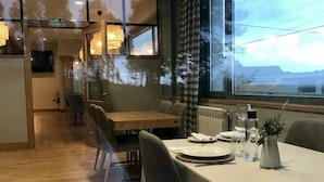 Een eetgedeelte met een tafel gedekt voor twee personen, uitzicht op het groen via een groot raam, en een keuken in de achtergrond.