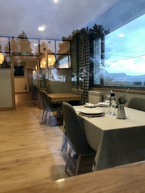 A dining area with a table set for two, a view of the outdoors through a large window, and a kitchen area in the background. - Hotel Restaurante Rias Altas (Barreiros)