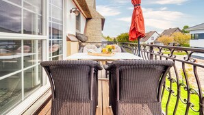 Een balkon met rieten meubilair, een tafel met fruit en bloemen, en een rode parasol.