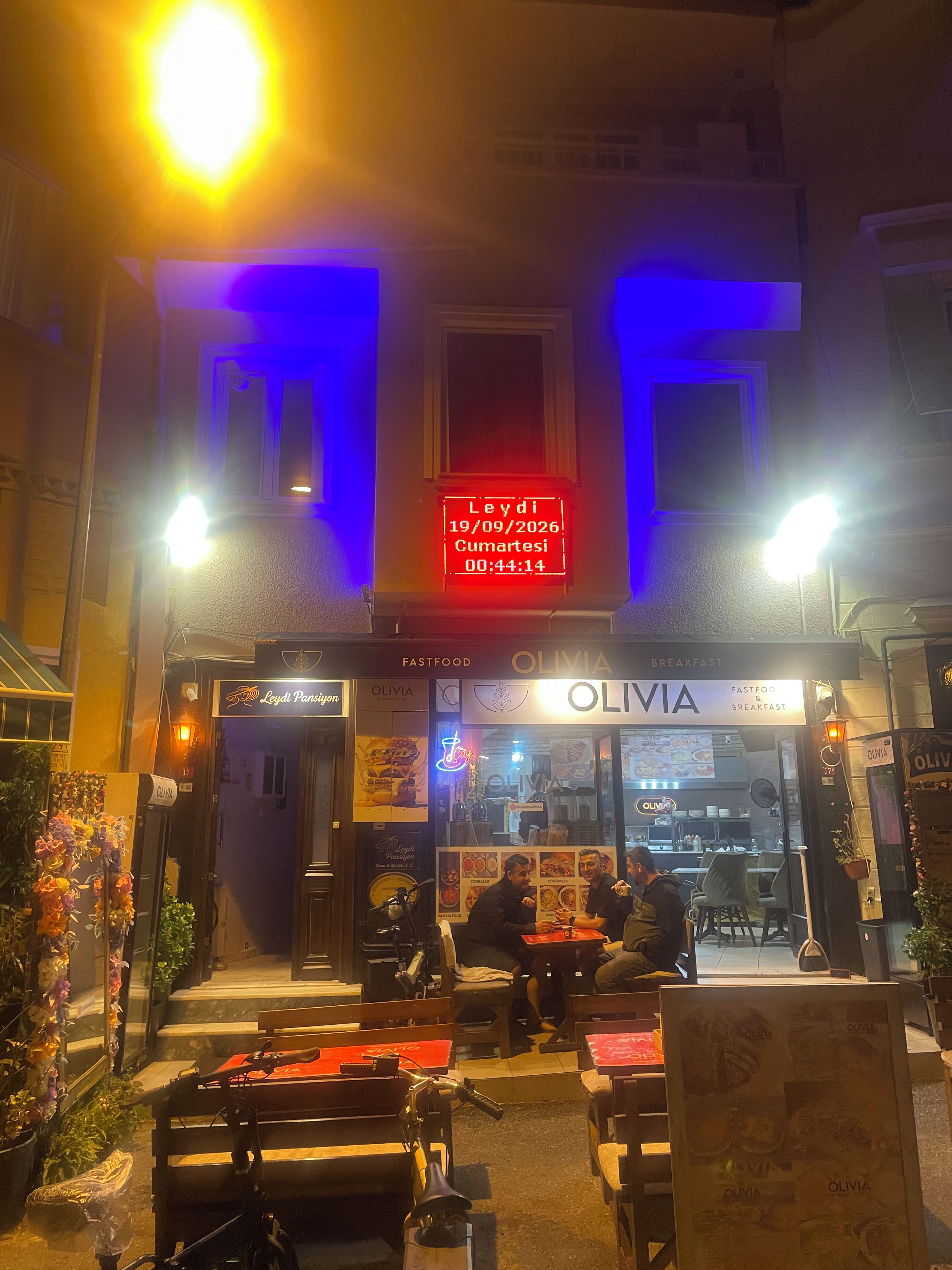 Ein Restaurant namens Olivia mit Außenbereich und einer Neonreklame, die Datum und Uhrzeit anzeigt.