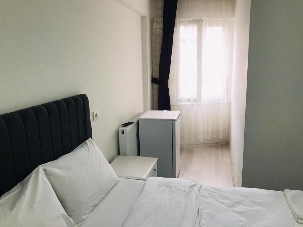 A hotel room with a bed, a bedside table, a small cabinet, and a window with sheer curtains.