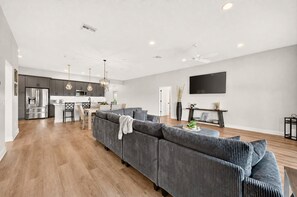A modern living room with a grey sofa, wooden flooring, and a kitchen in the background. - Fairway Vista - 3/3 w/ 4-Seat Golf Cart in The Villages - Moultrie Creek (Moultrie Creek, The Villages)