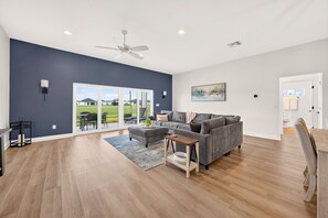 Ein geräumiges Wohnzimmer mit grauem Sofa, einem Holztisch und einem großen Fenster mit Blick auf eine grüne Landschaft.