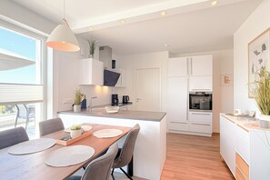 A modern kitchen with white cabinetry, a central island, and a dining area with a wooden table and chairs. - White Villas Penthouse 11 (Ostseebad Boltenhagen)