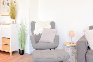 A modern living room with a grey armchair, ottoman, and a small side table with a lamp. There is a tall plant in a black pot and a framed picture on the wall. - White Villas Penthouse 11 (Ostseebad Boltenhagen)