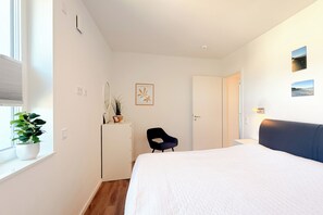 A bedroom with a bed, a desk, a chair, a potted plant, and a door leading to another room. - White Villas Penthouse 11 (Ostseebad Boltenhagen)