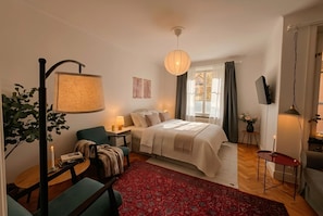 Ein helles Schlafzimmer mit Bett, Nachttischen, Schreibtisch, Stuhl, einem roten Teppich, einer Pendelleuchte, einem Fernseher und einem Fenster mit Vorhängen.