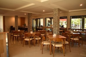 A spacious indoor dining area with multiple wooden tables and chairs, stone pillars, and large windows allowing natural light.