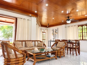 A room with a wooden ceiling, a glass coffee table, wicker furniture, and a dining area with chairs and a table.