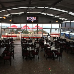 A restaurant with tables and chairs, a television screen, and large windows with umbrellas. - Norwegian Lodge (Umkomaas)