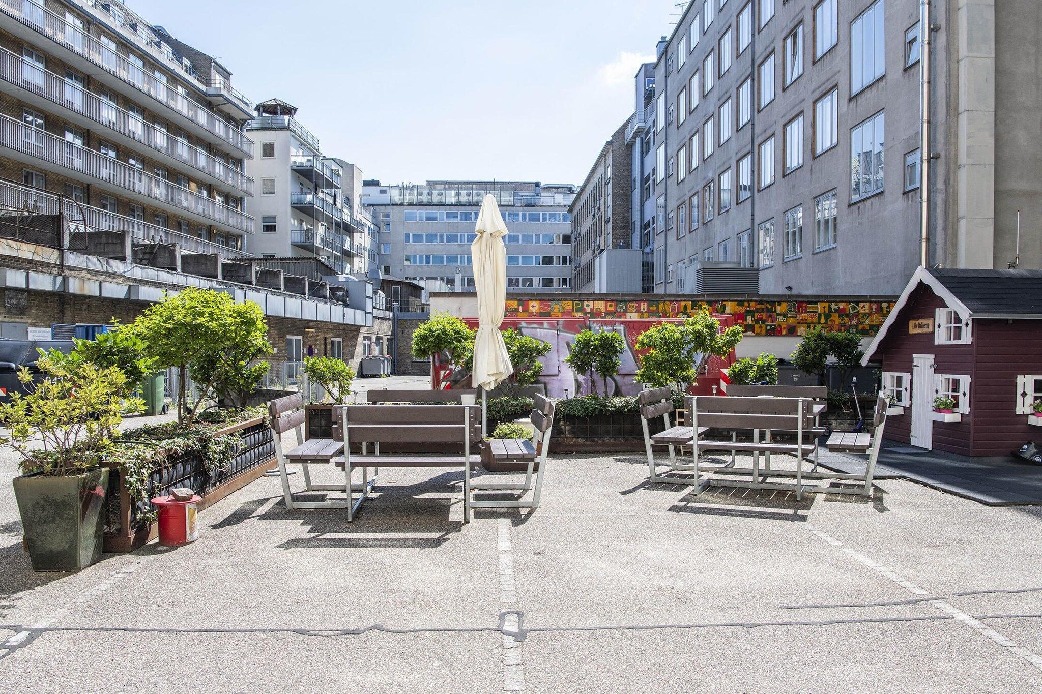 An outdoor seating area with benches and an umbrella, surrounded by buildings and greenery.