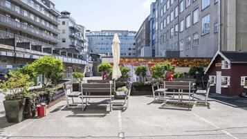An outdoor seating area with benches and an umbrella, surrounded by buildings and greenery.