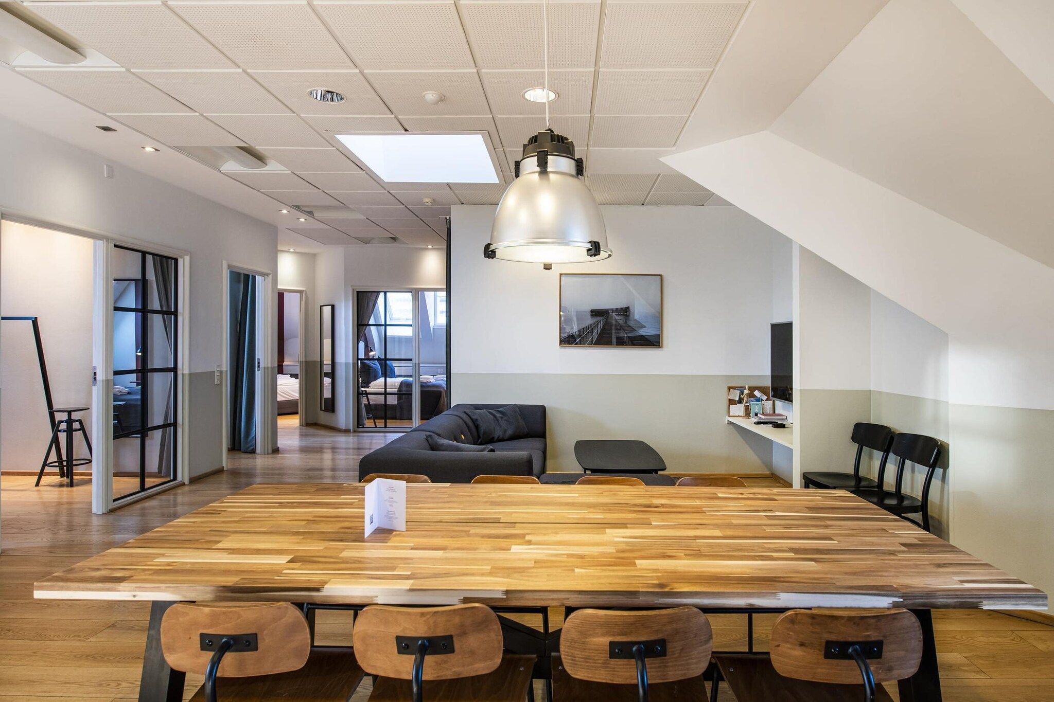 Et moderne kontor med et stort trækonferencelbord, sorte stole, en sort sofa og en hængende pendellampe.