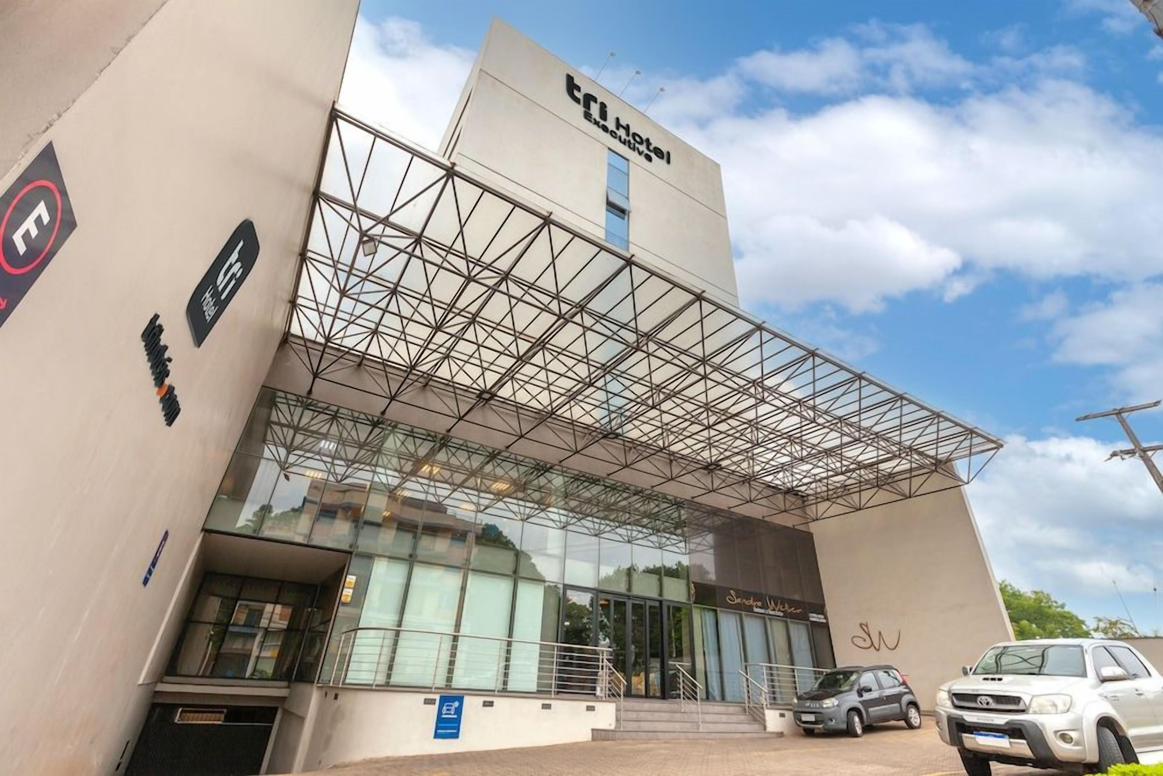 Um hotel moderno com fachada de vidro, cobertura em estrutura metálica e uma placa indicando ser um 'TRI Hotel Executive'.