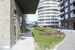 Edifício moderno com fachada de tijolos, estrutura de varanda circular e área externa pavimentada com um banco e vegetação.