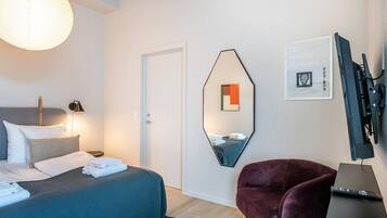 Ein Hotelzimmer mit einem Bett, einem Sessel, einem Schreibtisch mit Computer, einem Spiegel und einem an der Wand befestigten Fernseher.