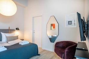 Quarto de hotel com cama, poltrona, escrivaninha com computador, espelho e televisão na parede.