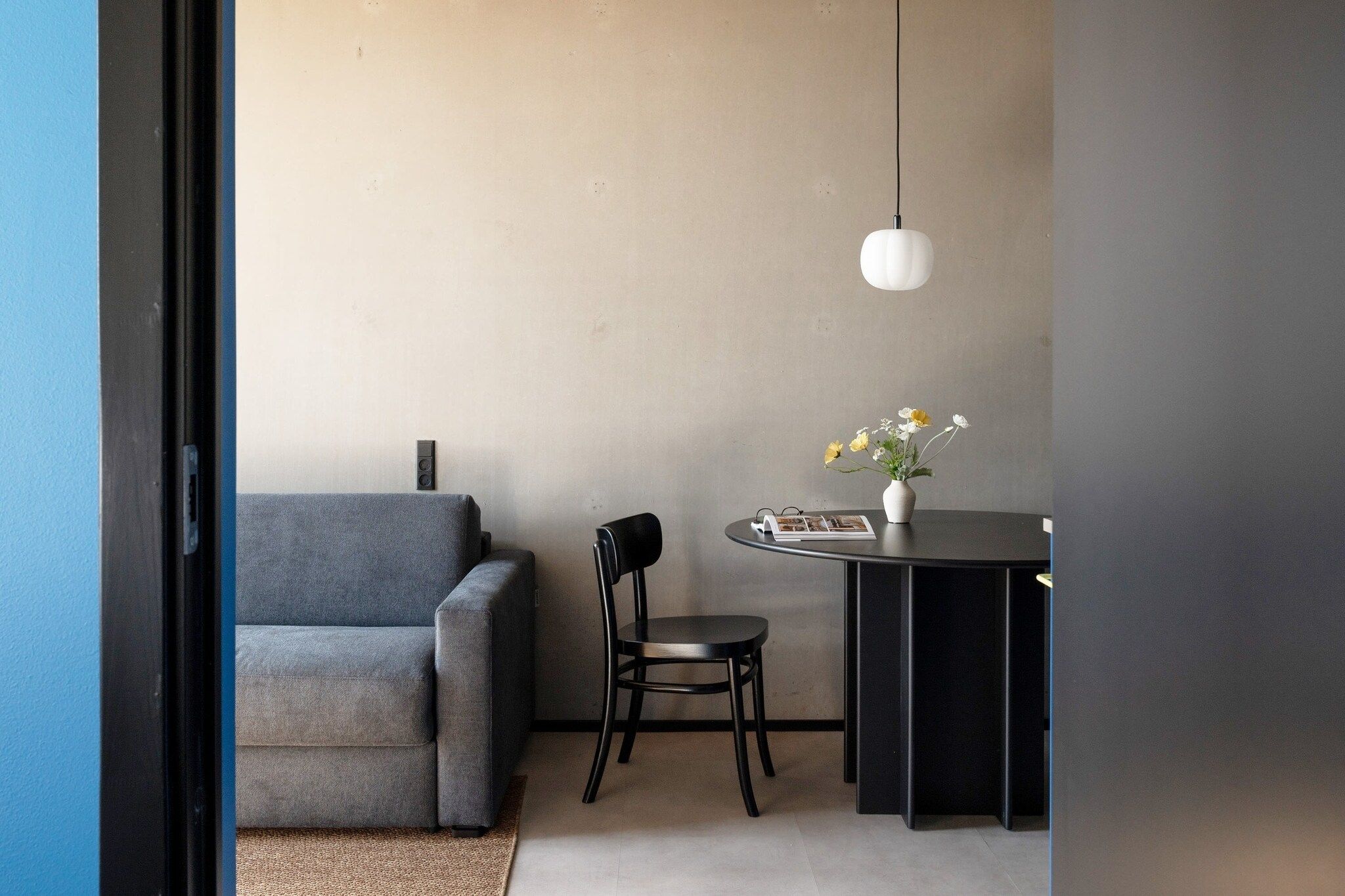 A room with a grey sofa, a black chair, a small round table with a vase of flowers, and a hanging light fixture.