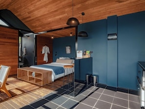 A modern bedroom with a wooden ceiling, a bed with a blue bedspread, a wooden bench, and a small table. - The Boato Hotel (El Peñol)