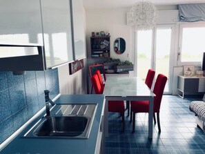 A modern kitchen with a built-in sink, a dining area with red chairs, and a view of the outdoors. - Seafront Apartment by the City Centre (Lignano Sabbiadoro)