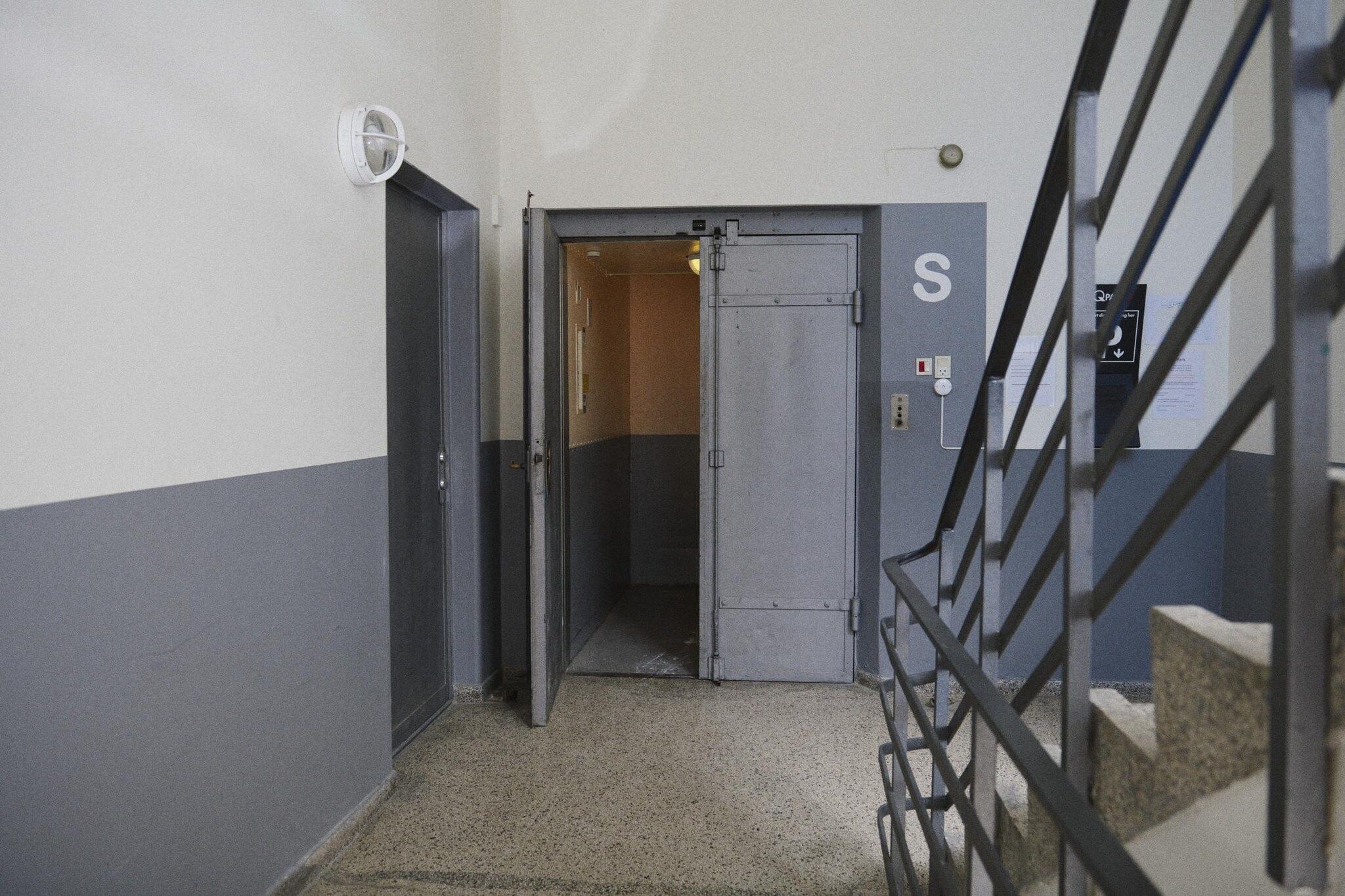 Un couloir étroit avec une porte marquée d’un panneau « S », un escalier en métal et un éclairage de sécurité fixé au mur.