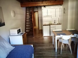 Habitación compacta con cama, una kitchenette pequeña, una mesa de madera con sillas y una escalera que lleva a una zona elevada.