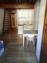A narrow hallway with a wooden staircase, a kitchen area with white cabinets, and a dining table with chairs.