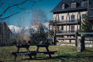 Una mesa de picnic de madera en un área con césped, con un edificio de piedra y montañas al fondo.