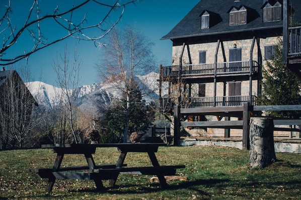 Una mesa de picnic de madera en un área con césped, con un edificio de piedra y montañas al fondo.