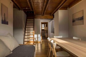 Un interior acogedor con una escalera de madera, un comedor con una mesa larga y sillas, y una cama con una manta gris.