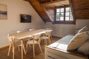 Una habitación con una mesa y sillas de madera, una cama con almohadas, un televisor montado en la pared y un ventanal con vista a árboles.