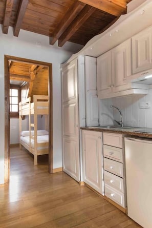 Una cocina con armarios blancos, techo de madera y una litera en la habitación contigua.