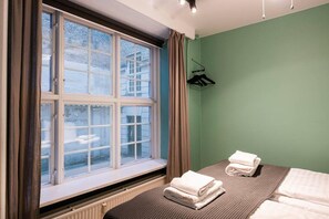 Een hotelkamer met een groot raam, een bed met twee kussens, een radiator en een hangende kapstok.