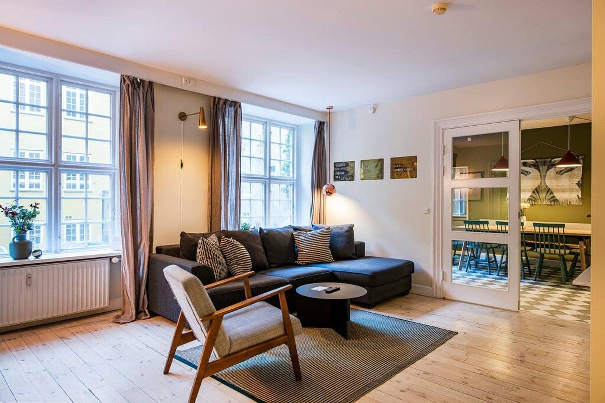 A modern living room with a sofa, armchair, and coffee table, large windows with curtains, and a view of the city.
