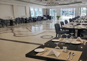 A hotel dining area with tables set for service, a chandelier, and a view of the street through large windows. - Carawan Al Khaleej Hotel Olaya (Riyadh)