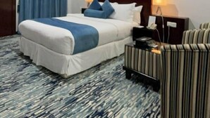 A hotel room with a large bed, a bedside table with a lamp, a striped armchair, and a painting on the wall. - Carawan Al Khaleej Hotel Olaya (Riyadh)