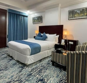 A hotel room with a large bed, a bedside table with a lamp, a striped armchair, and a painting on the wall. - Carawan Al Khaleej Hotel Olaya (Riyadh)