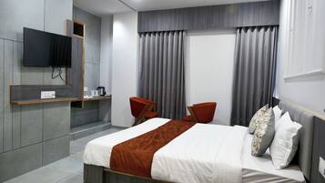 Quarto de hotel moderno com cama, TV fixada na parede, pequena mesa com cafeteira e um espaço de estar com duas cadeiras.