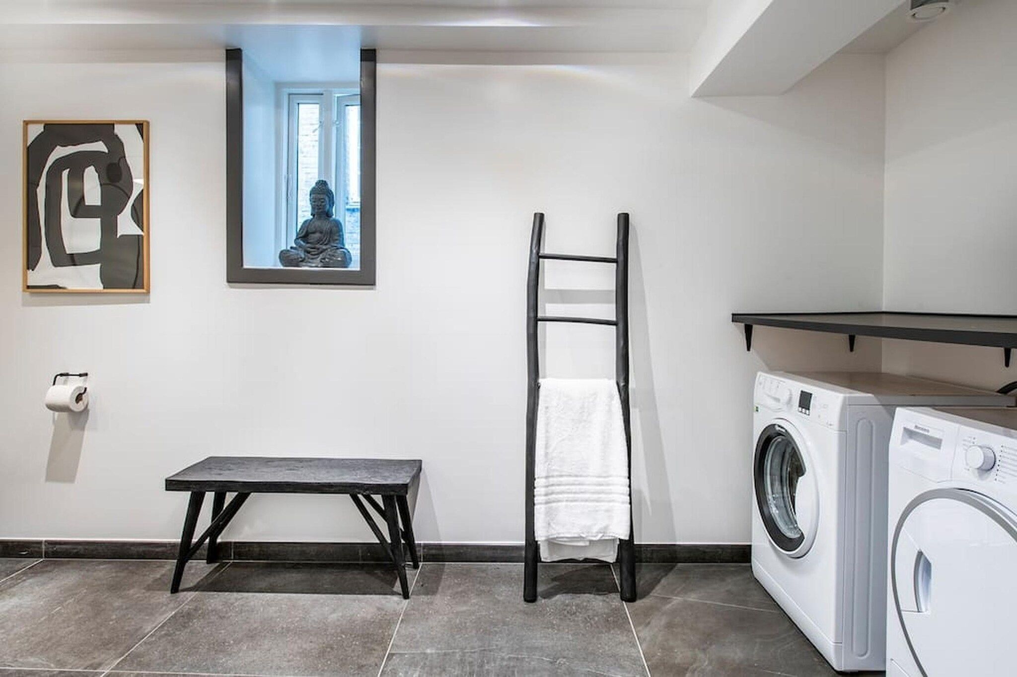 A laundry room with a washing machine, a drying rack, a bench, and a framed picture on the wall.