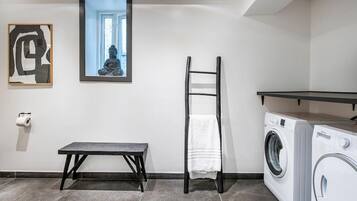 A laundry room with a washing machine, a drying rack, a bench, and a framed picture on the wall.